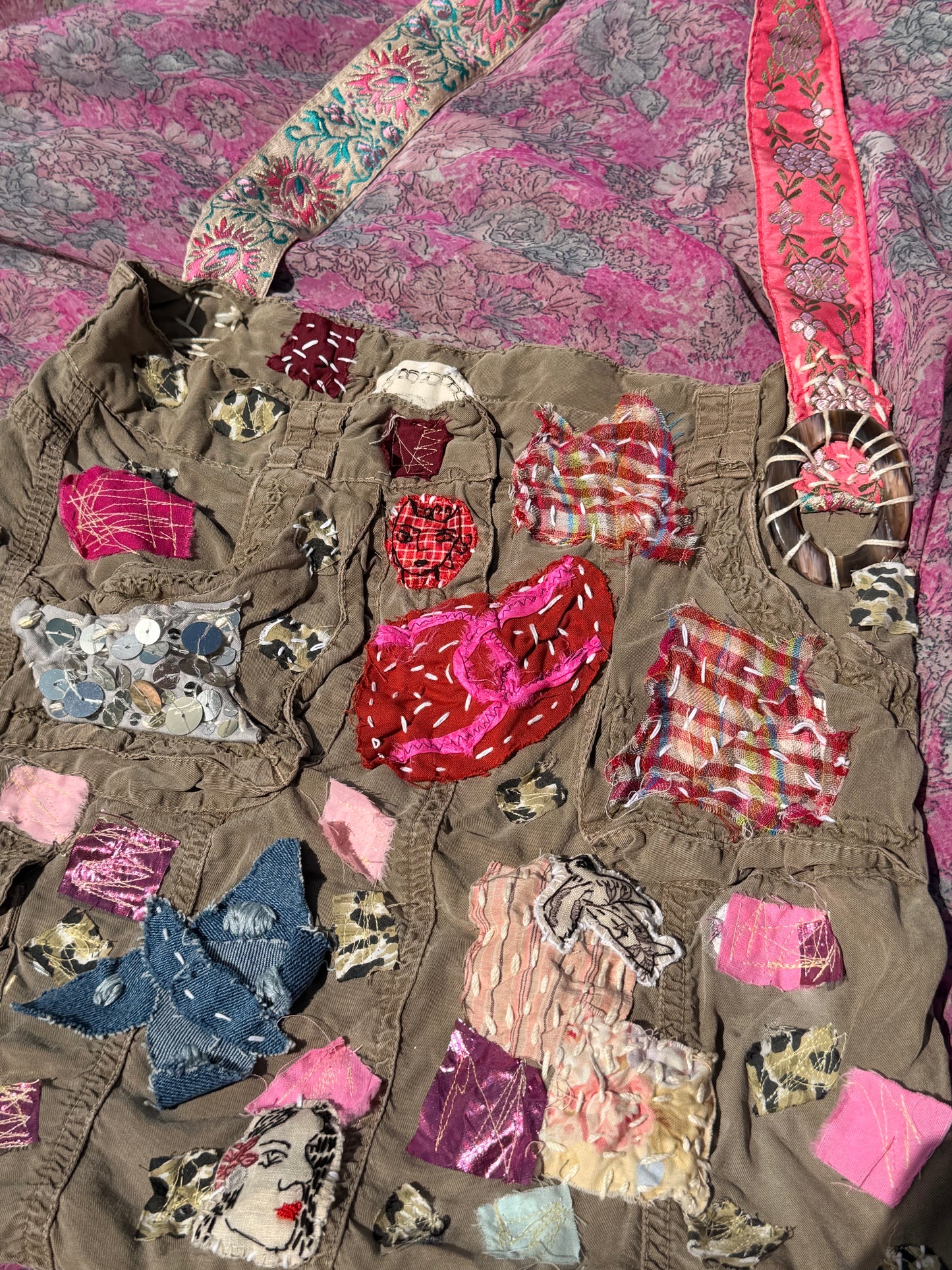 A handmade brown patchwork vest with multiple colorful fabric pockets and appliqués, laid flat on a pink patterned fabric background.